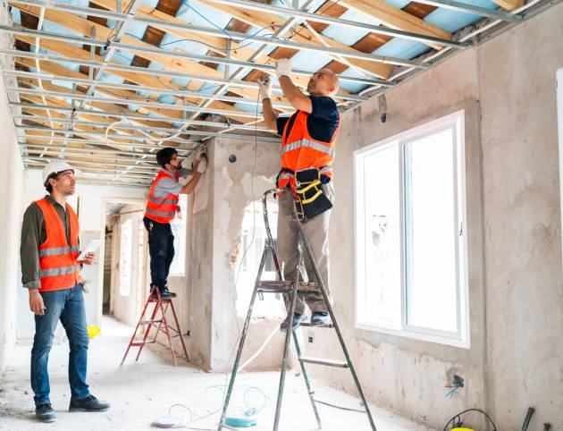 Three construction workers in safety vests and helmets work together installing ceiling framework and windows during an interior renovation project.